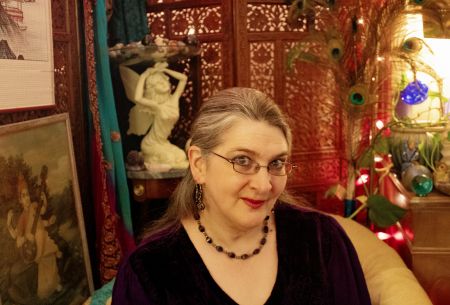 A gently smiling woman wearing dark purple sits before a carved teak screen from India and a statue of a kneeling fairy.  A picture of the Hindu goddess Sarasvati appears next to her, on the viewer's left.  Pink lights twinkle next to peacock feathers on the viewer's right.  